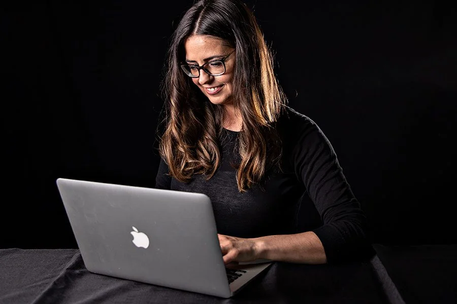 Girl with brown hair sitting in front of a grey apple laptop giving virtual help on a black backdrop